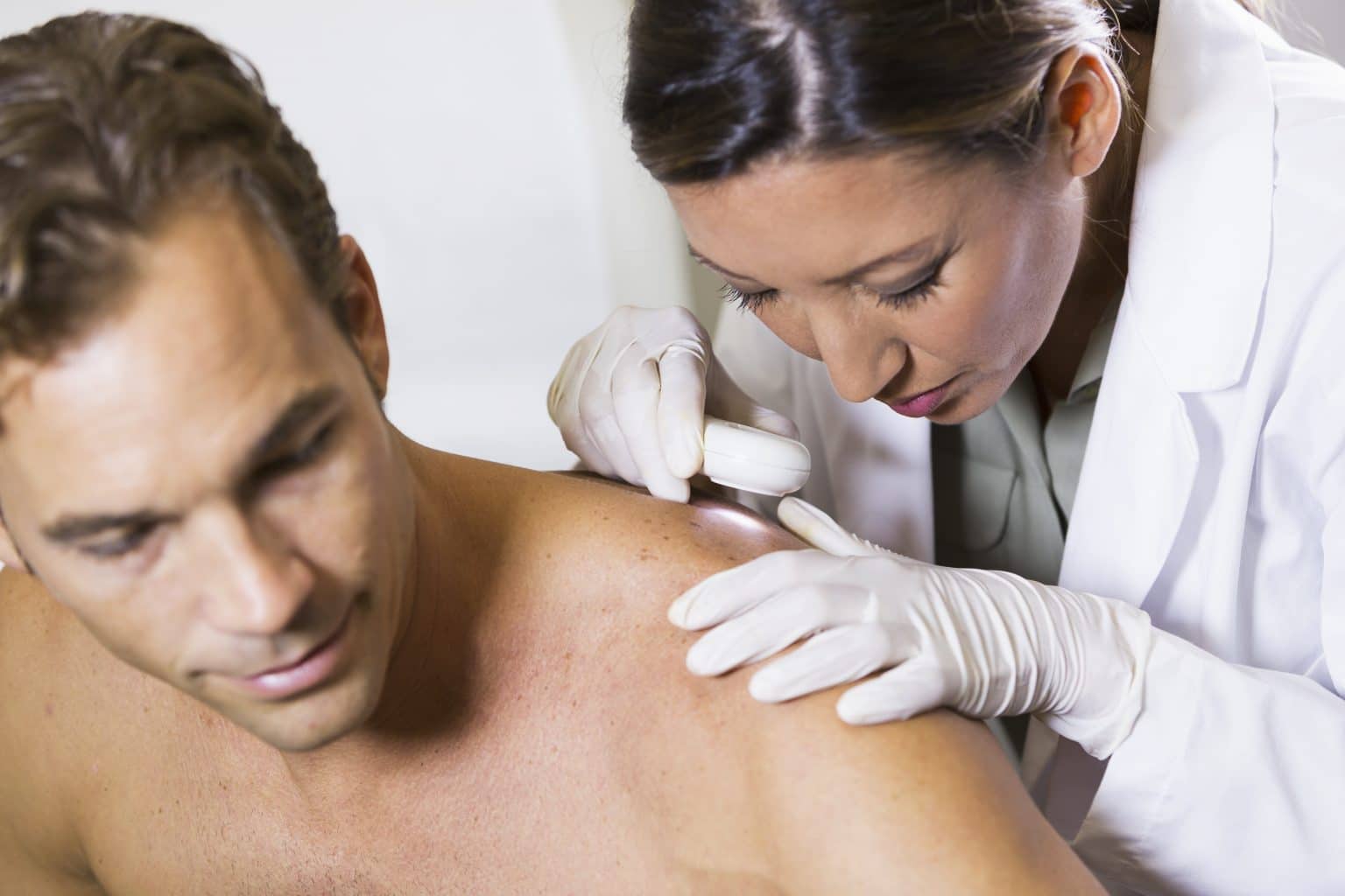 Female physician examining male patient with dermascope, looking for signs of skin cancer.